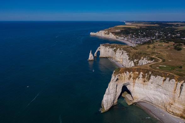 Aerial view of white chalk cliffs along a coastline with a blue ocean, featuring distinctive rock formations and a small beach area. In the background, a town is visible.