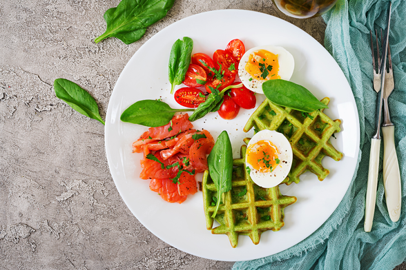 Plato con gofres salados, salmón ahumado, espinacas, huevos cocidos y tomates cherry sobre mesa de piedra gris.