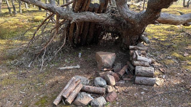 A few logs leaned up against a tree making a shelter that looks like they might fall on someone who sits on the rock inside. 
Trap baited with a fireplace infront and firewood on the side and a beercan lying deep inside the shelter.