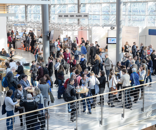 Blick von oben in die Kongresshalle. Kongressbesucher*innen stehen an Stehtischen und Ausstellungsständen.