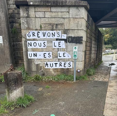 Collage de lettres blanches sur le pilier d’un pont qui dit : Grevons nous les un•es les autres.