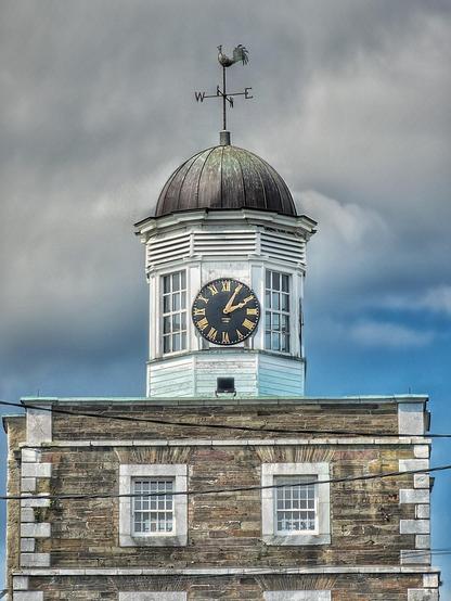 A close-up shot of the Youghal Clock Gate tower. The weathered brick building supports a white octagonal lantern, which houses a large black clock face with Roman numerals and gold hands. A weathervane with a cockerel sits atop the dark, domed roof. The sky behind is cloudy with patches of blue.