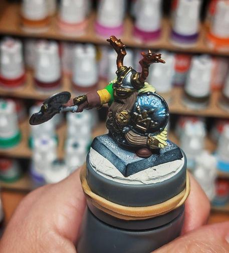 Figura de un enano de fantasía pintada a mano. Lleva una armadura de tonos grises y dorados y un casco con cuernos. El enano tiene una barba larga y marrón y sostiene un escudo circular con un diseño brillante y un hacha de batalla de doble filo. La figura se apoya sobre una peana de forma cuadrada. El fondo de la imagen está desenfocado y se aprecian filas de botes de pintura de diferentes colores, indicando que la foto se ha tomado en un lugar donde se realizan manualidades o pintura de miniaturas.