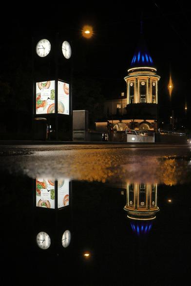 Das Bild zeigt eine nächtliche Straßenszene in einer urbanen Umgebung. Rechts steht ein markanter Turm eines historischen Gebäudes mit einer kuppelartigen Spitze, die in einem intensiven Blauton beleuchtet ist. Links befindet sich ein "Uhrturm" mit  beleuchteten Werbetafeln, auf denen verschiedene Speisen abgebildet sind. Die Straße ist nass, vermutlich durch kürzlichen Regen, wodurch sich die Szene deutlich in einer Pfütze auf dem Asphalt spiegelt. Diese Spiegelungen erzeugen eine fast symmetrische Wirkung im unteren Bildbereich. Der Himmel ist dunkel, und es sind keine Menschen oder Fahrzeuge sichtbar.