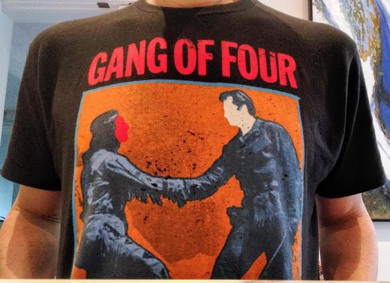 Close up photo of the front of my Gang of Four black t-shirt. The band name is written in red across the top. The image is taken from their 'Entertainment' record. Against a square orange background, there is an image of a cowboy (face is painted white) and a First Nations man (face is painted red) shaking hand. The album art was a comment on how colonizers exploited their relationships with First Nations peoples.