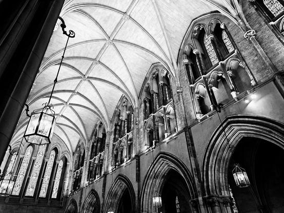 A dramatic black and white, low-angle shot looking up at the vaulted ceiling and arched windows of a gothic cathedral. The image highlights the intricate stonework and detailed patterns of the ceiling. Large, ornate light fixtures hang from the ceiling, and light streams through the tall, stained-glass windows, creating a sense of grandeur and historical scale.