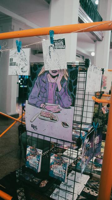Fotografía. Planta inferior del Antiguo Palacio Federal en el centro de Monterrey (México). Acercamiento al lateral de un stand en "Otro Bazar", parte de las actividades de Sábado en LabNL. Un par de zines cuelgan de un mecate mientras un poster cuelga de un rack y un poco más abajo se encuentran un par de ejemplares del xine con información general de Leyendas del Noreste, la comunidad de creación de juegos de rol del LabNL.