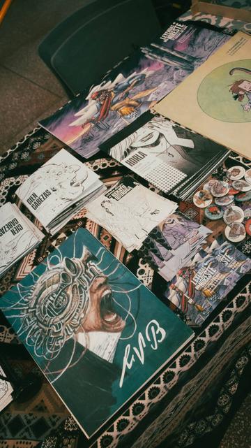 Fotografía mostrando la parte superior de una mesa con publicaciones (tres autopublicaciones) y otros productos como prints, pines y stickers de mi producción personal y de mi trabajo para Leyendas del Noreste, la comunidad de creación de juegos de rol.