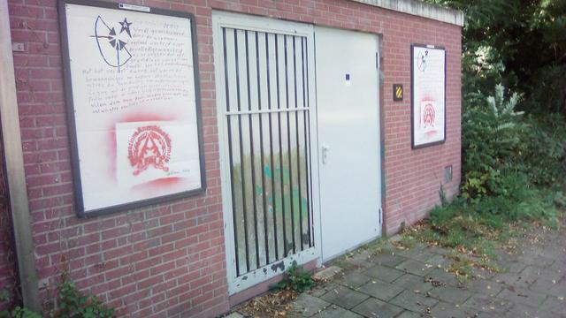 bakstenen muur met in het midden twee stalen deuren, lijkt het, waarvan 1 wit en de ander met spijlen. Aan beide zijden zijn de reclame borden bewerkt in, zie caption en alt in andere foto's. Links naast de muur groen begroeiing
