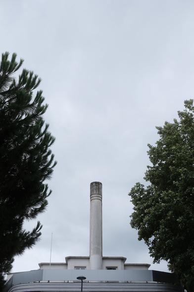 Photo au format portrait d’une cheminée au sommet d’un batiment assez petit, et au milieu du toit, donnant une symétrie au cliché. Des feuillages d’arbres viennent cadrer l’ensemble à gauche et à droite