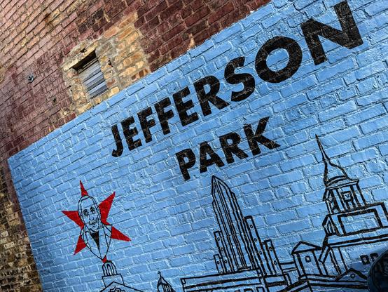 Mural painted on a brick wall featuring the words “JEFFERSON PARK” in bold black letters on a light blue background. To the right of the text is a red star with a stylized black-and-white portrait of Elijah Wentworth, depicted with a stern expression. Below the text and portrait are black line drawings of Chicago architecture, including a skyscraper and a steepled building, representing local landmarks. The mural celebrates the history and identity of Jefferson Park, a neighborhood in Chicago.