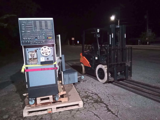 a palletized univac 418, teletype desk on its right, as seen from the front panel,  in a parking lot. a forklift is parked to the right of the whole ordeal.
