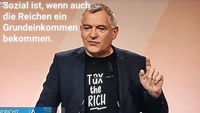 Jan van Aken mit erhobenem Zeigefinger, auf seinem T-shirt steht tax the rich, im Hintergrund spruch: sozial ist, wenn auch die Reichen ein Grundeinkommen bekommen.
