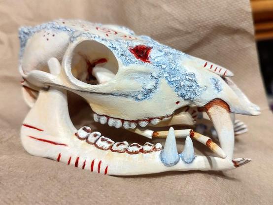 Side view of a creature skull, deer-like with a prominent fang on the upper jaw, and two fangs on the lower. It also has antennae and small hands inside the mouth. The skull is tan color with blue-ish spots and red carved patterns.