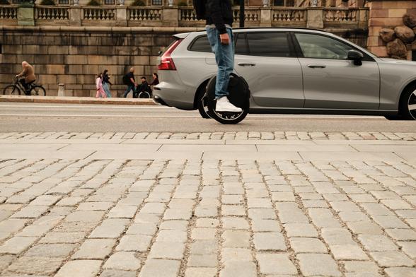 A person rides an electric unicycle on a cobblestone street next to a silver car, while cyclists and pedestrians pass by in the background near a stone wall. The unicycles wheel is in the same position as the car's back wheel.