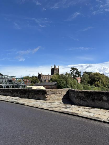 Photo taken on a bridge looking towards Hereford Cathedral in the distance. The sky is blue.