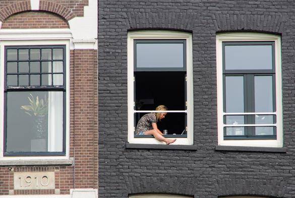A picture of windows upstairs on a street in Amsterdam. Two buildings, red brick to left with one window and a date of 1910, the right hand building is brick painted black, two windows, and a women is sitting inside the window, possibly typing on a laptop, one leg is out the open window over the window ledge, probably to keep cool.