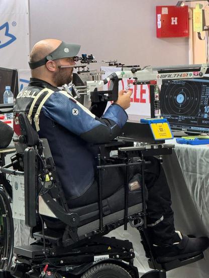 A man in a wheelchair aims an air rifle on an indoor shooting range. A metal stand takes the weight of the rifle, leaving him to aim and release the shot.