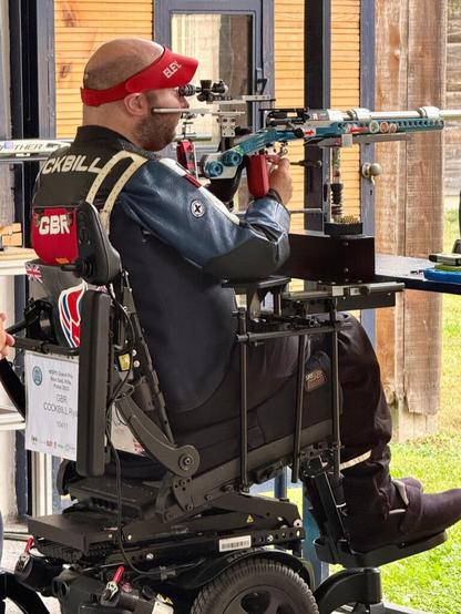 A man in a wheelchair aims a .22lr rifle on an outdoor shooting range. A metal stand takes the weight of the rifle, leaving him to aim and release the shot.
