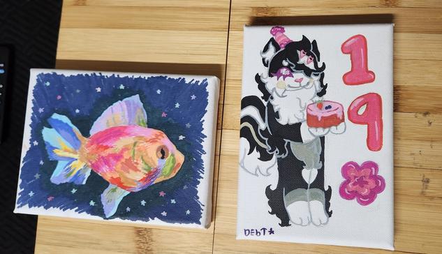 Two canvas paintings side by side. The one on the left is a brightly colored fish swiming in a dark space. The one on the right is an anthro skunk holding a birthday cake, the number 19 is printed on the side with a large flower below it.