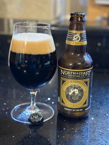 A snifter full of dark beer with a thick light tan head. The short brown bottle has a black and gold label with a black and white drawing of Rasputin with his fingers to his forehead.
