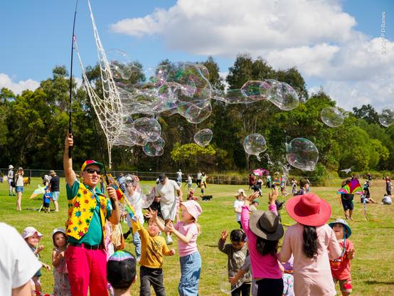A children's entertainer standing in a large grassed field holds a large mesh bubble-make aloft as dozens of large bubbles stream out of it to the obvious delight of children gathered around.