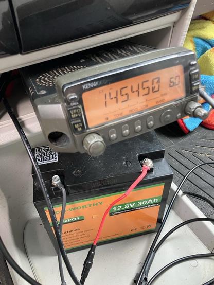 An old Kenwood TM-G707A dual band radio is crammed into a small cavity in a vehicle's middle console. Under that, a 12.V 30Ah Eco-Worthy LiFePo4 battery sits on the floor board. Wires connect things and extend beyond the photo's borders.