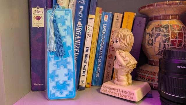 A bookshelf with several books, including "Mom’s Devotional Bible," "The Great Gatsby," and "Linda Goodman’s Sun Signs." In front of the books is a blue bookmark decorated with puzzle piece patterns and a shiny blue tassel. Beside it sits a small ceramic figurine of a blonde child holding a heart that says “I Love You,” standing on a base that reads, “Sometimes I Hate You but Always I Love You.” A mosaic vase and a camera lens are also partially visible.