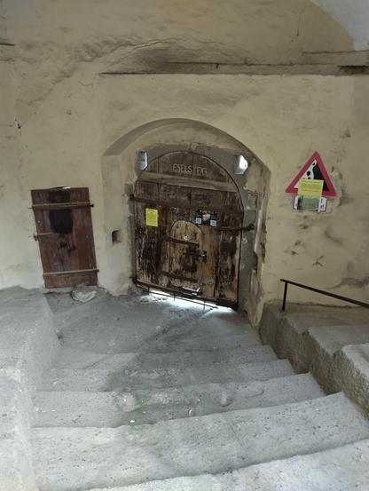 photo depicts a small door and a gate, both made of old wood in a plastered stone wall, down steep stone stairs, the word "Eselsteig" carved into the gate