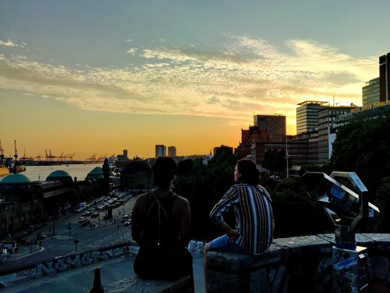 Zwei mir unbekannte Menschen genießen den Ausblick am Abend eines lauen Tages  auf der Umrandungsmauer des Aussichtspunktes oberhalb der Hamburger Landungsbrücken. Der Blick geht über das Gebäude der Landungsbrücken Richtung Nordsee über den Hafen im Sonnenuntergang