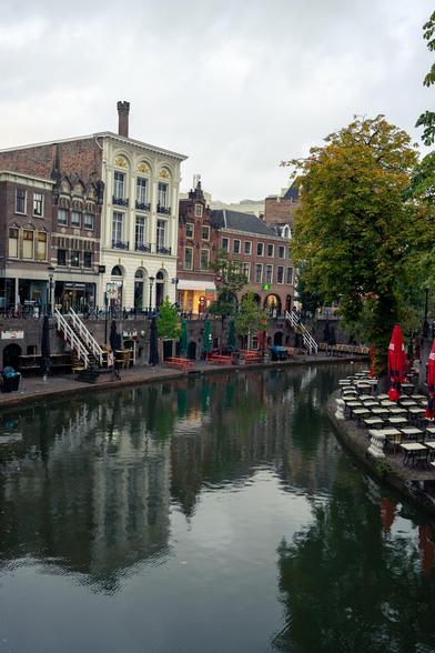 "Image of a picturesque canal scene in a European city featuring elegant buildings with varied architectural styles lining both sides. The buildings exhibit ornate facades, large windows, and a mix of brick and white exteriors. On the right side of the canal, there is outdoor seating with tables, chairs, and large red umbrellas. The calm canal water reflects the buildings and surrounding green trees, contributing to the serene atmosphere. A few benches and a person visible on the left side add to the human element in the scene."