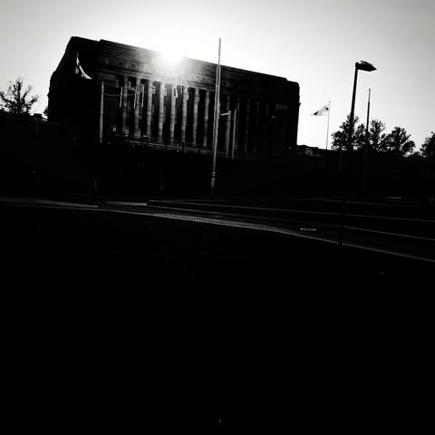Silhouette of the Finnish Parliament House against a bright sun, surrounded by trees and flags, creating a dramatic black-and-white scene.