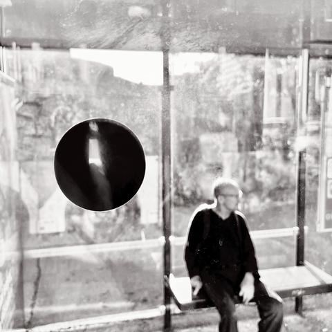 A person sits at a brightly lit bus stop, framed through a bus window with a large black circle on the glass, creating a contrast against the blurred background.