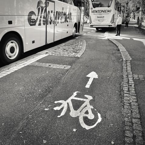 A bike lane marked with a bike symbol and directional arrow, adjacent to two parked buses effectively blocking the bike lane, in a monochrome city setting.