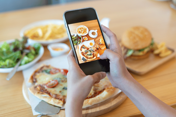 Persona tomando foto de pizza con un smartphone; en segundo plano, hamburguesa, patatas fritas y ensalada sobre una mesa de madera.