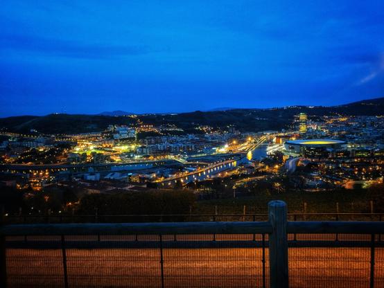 A long-exposure, horizontal shot of a city at night. The city's lights illuminate the buildings, roads, and a large stadium in the foreground. A river runs through the centre of the city, crossed by several lit bridges. The surrounding hills are dotted with lights, adding to the urban glow. The sky is a deep blue, characteristic of the 'blue hour' after sunset. A dark railing and a post are visible in the foreground, framing the vibrant city below. The overall image gives a sense of a bustling, active city.