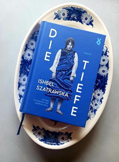 Foto des Buches, auf dessen blauem Cover eine junge Frau mit traditioneller blauer Kleidung und Haube steht. Sie lässt die Arme hängen und schaut von unten in die Kamera. Das Buch liegt auf einer mit blauen Blumen bemalten weißen Servierplatte, die auf grauem Hintergrund steht. Das Foto wird von oben nach unten dunkler.