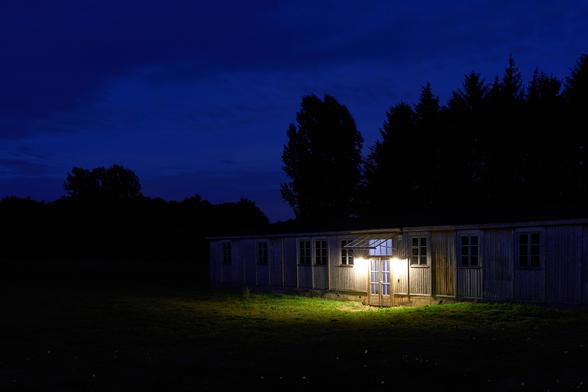Nachtaufnahme einer Baracke auf dem Gelände der Gedenkstätte. Die Aufnahme wurde nach Sonnenuntergang gemacht und damit ist der Himmel dunkelblau. Erkennbar ist noch der Eingang einer Baracke. Ein Oberlicht beleuchtet die Tür des Gebäudes.