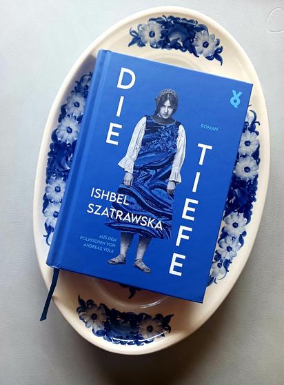 Foto des Buches, auf dessen blauem Cover eine junge Frau mit traditioneller blauer Kleidung und Haube steht. Sie lässt die Arme hängen und schaut von unten in die Kamera. Das Buch liegt auf einer mit blauen Blumen bemalten weißen Servierplatte, die auf grauem Hintergrund steht. Das Foto wird von oben nach unten dunkler.