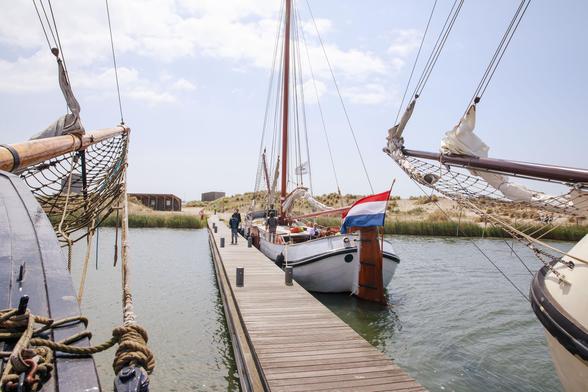 Aanlegsteiger bij een eiland van de Marker Wadden. Op de voorgrond zie je de boeg van twee boten en, daarachter volledig in beeld, een zeilschip dat aan de steiger ligt. Op de achtergrond zie je duingebied en enkele houten huisjes.