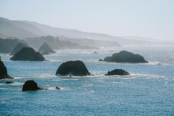 A coastal view with distant mountains, rocky formations in the ocean, and gentle waves under a hazy blue sky.