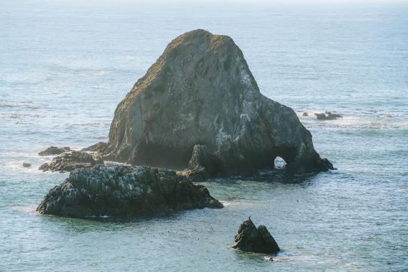 A prominent rock formation stands in the ocean, surrounded by smaller rocks, with calm waters reflecting the light.