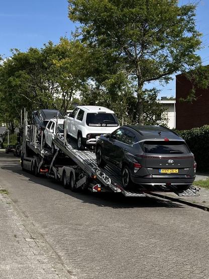 Vier nieuwe auto’s die op een vrachtwagen staan in een straat. Op de achtergrond groene bomen en blauwe lucht.