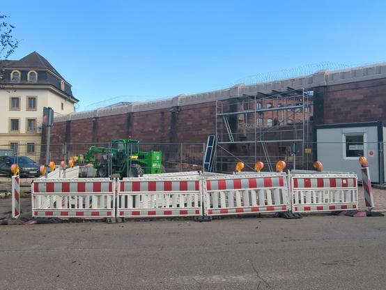 Blick auf eine Gefängnismauer von außen. In der Bildmitte befindet sich ein Baugerüst an der Mauer und in der Mittes des Gerüsts sieht man ein großes Loch, da ein Bauarbeiter die Mauer einreißt. Links von dem Gerüst steht ein grün farbener Bagger. Durch das Loch in der Mauer hindurch, sieht man vergitterte Zellenfenster. Ganz links im Bild ist noch ein Teil eines Gefängnisbaus, vierstöckig, mit vergitterten Fenster zu sehen.