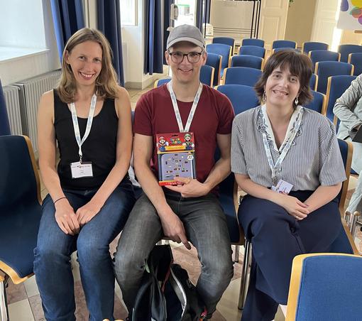 Photo of the three members of “Team Baby Mac” after winning the Open Knowledge Maps Scientific Scavenger Hunt at the Open Science Festival 2025. They are sitting next to each other on chairs in one of the event’s locations. The person in the centre holds up a prize.