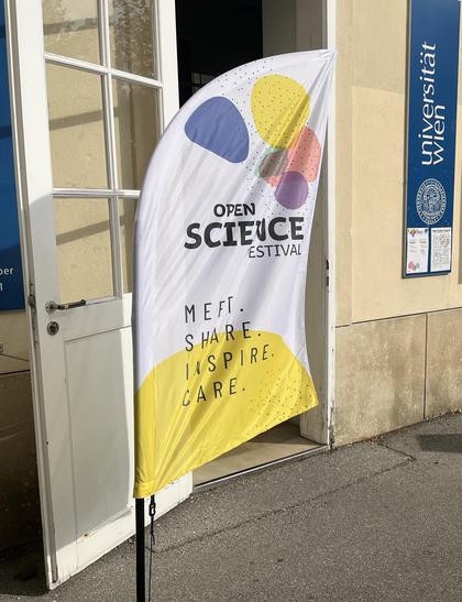 A flag for the Open Science Festival 2025 stands outside the entrance to the Aula at the University of Vienna campus. The flag features the festival’s keywords “Meet”, “Share”, “Inspire”, and “Care”.