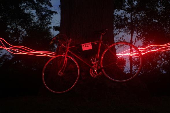 light-painting photo of my classic road bike leaning against a large tree at night; it's lit with an otherworldly red glow, and thin, squiggly streaks of bright red light arc behind the tree and off each side of the image