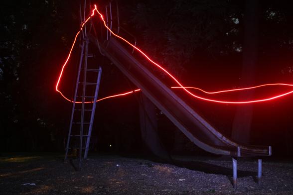 light painting photo of a metal slide at night; the slide and the area around it are lit by a cool white light coming from the general direction of the camera, while a single bright red streak of light trails in from the right edge of the frame, up the ladder, down the slide, and disappears again off the right side
