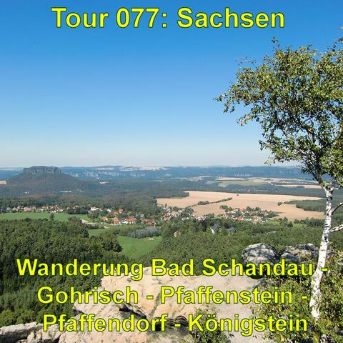 Das Bild bietet einen weiten Blick über eine hügelige Landschaft. Im Vordergrund ist der Rand eines Felsplateaus zu sehen, das dicht mit Bäumen bewachsen ist, darunter auch ein hervorstechender Baum mit grünen Blättern auf der rechten Seite. In der Ferne erstrecken sich Felder und Wälder, unterbrochen von einem kleinen Dorf mit zahlreichen Häusern. Im Hintergrund erheben sich weitere bewaldete Hügel, und der blaue Himmel ist weitgehend klar mit nur wenigen Wolken. Die Szenerie wirkt ruhig und malerisch und ist typisch für ländliche Gegenden.