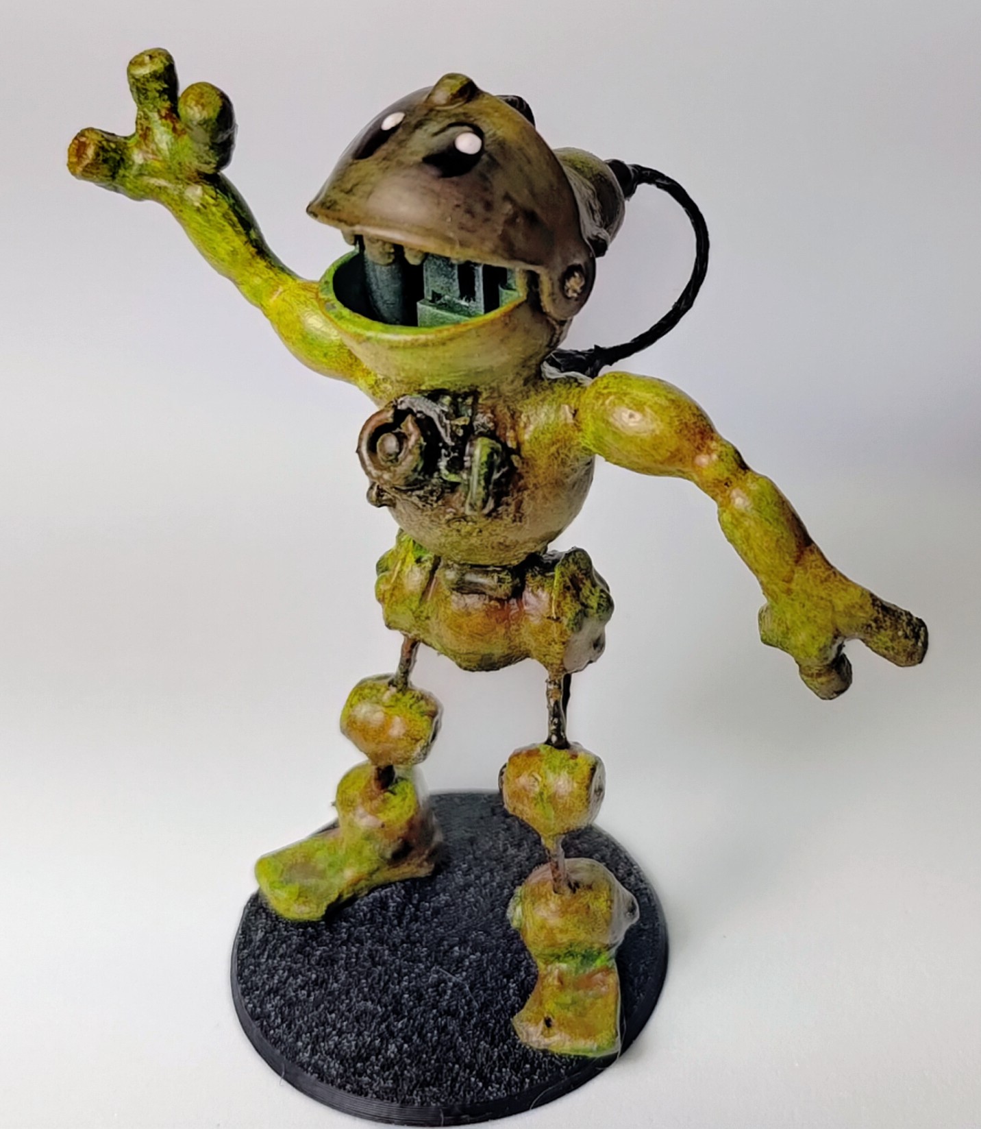 A miniature robot whose head is made with one of the toy fish. He's green and bronze and looks kind of froggy. He has three fingers and kind of organic arms, his torso has some mechanical bits and his legs are mechanical joints and feet with metal bars connecting them. His head has a big wire coming out the back of it and connecting between his shoulder blades. He still has cartoon eyes and his waving with his mouth open and happy.